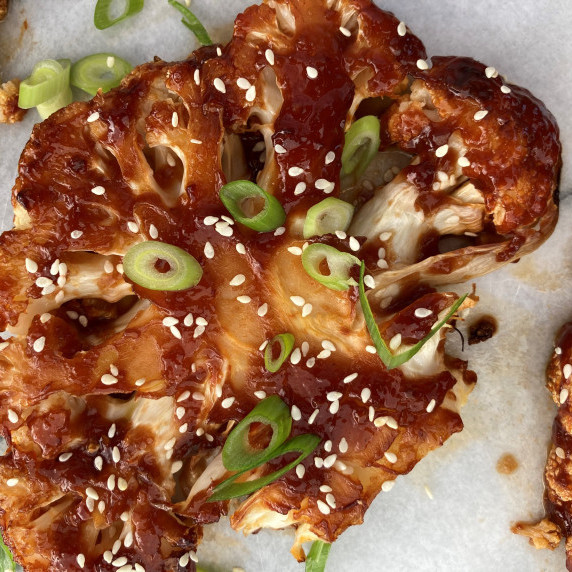 One glazed cauliflower steak topped with sliced scallions and sesame seeds on a marble board. 