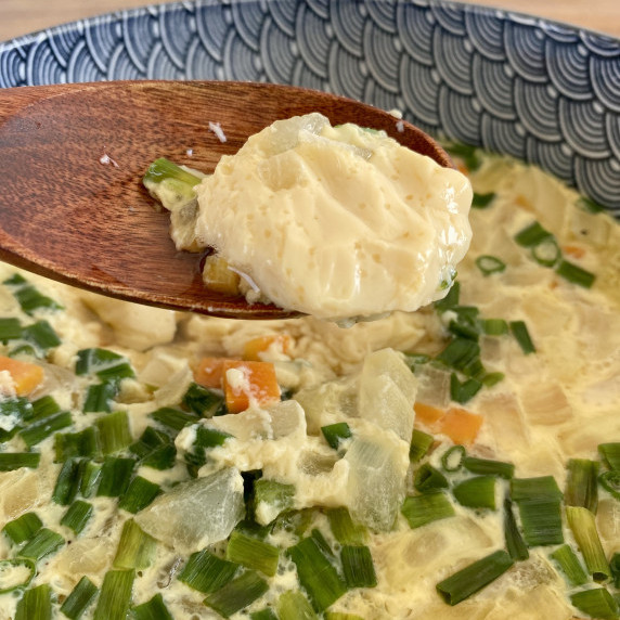 A wooden spoon holding steamed egg above a bowl filled with Thai steamed egg in it.