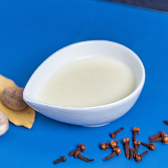 bechamel sauce in a small white bowl with cloves, nutmeg, and bay leaves surrounding it