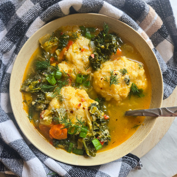 A bowl filled with golden broth, pops of green, & dumplings against a checkered towel