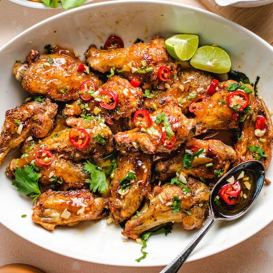 Air fried chicken wings with sauce, red chili peppers, cilantro and lime in a white bowl with spoon