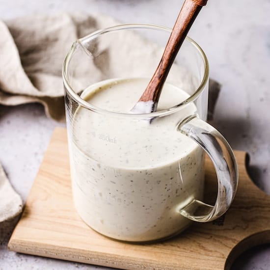 Ranch dressing in a glass pitcher with a wooden spoon atop a cutting board