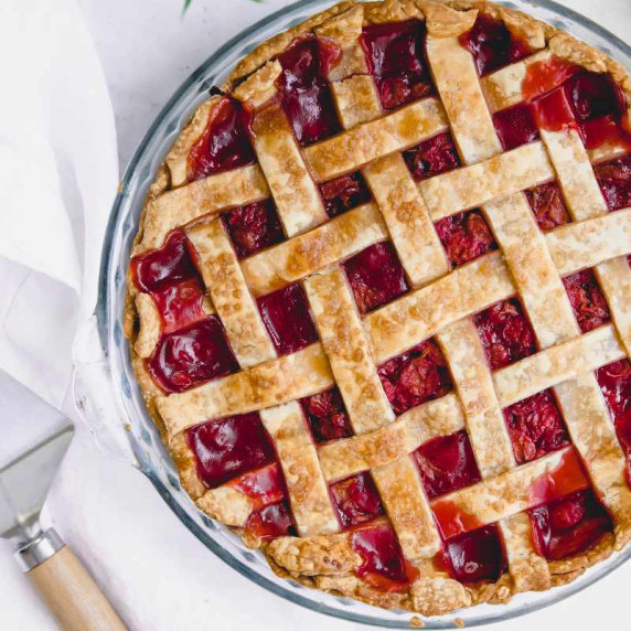 A whole cherry pie with a lattice top in a glass pie plate rests with a pie server beside it.