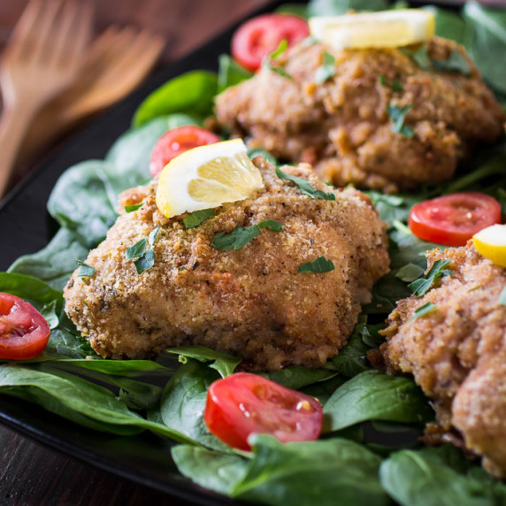 Side shot of multiple pieces of breaded chicken garnished with herbs and a lemon wedge on spinach.