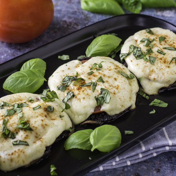 Side shot of portabella mushrooms garnished with basil on a black rectangle plate with a tomato in t