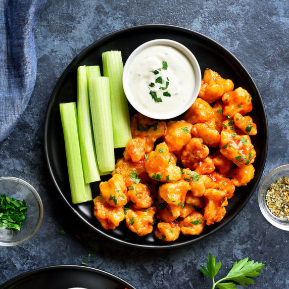 Cauliflower in orange paprika on a black plate with green celery and white ranch dressing on a dark 