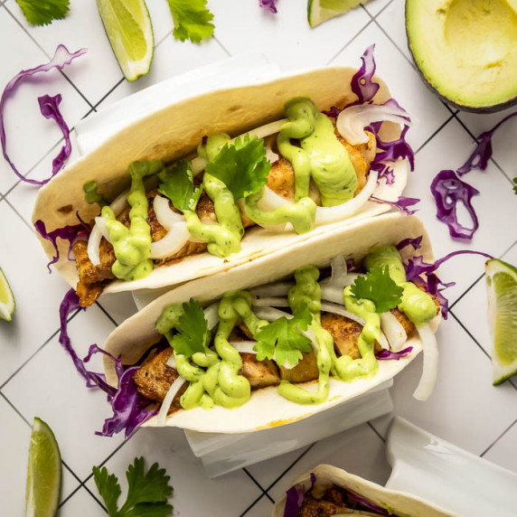 Overhead shot of two tacos with avocado cilantro sauce and fresh cilantro on top on a tiled surface.