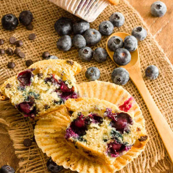 A blueberry chocolate chip muffin cut in half on a paper liner on burlap next to blueberries.