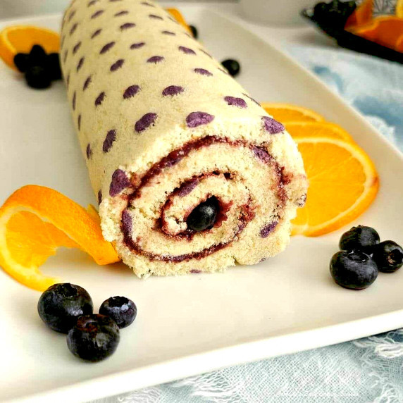 End shot of a purple dot patterned Swiss roll cake, with bluebrries on white platter.