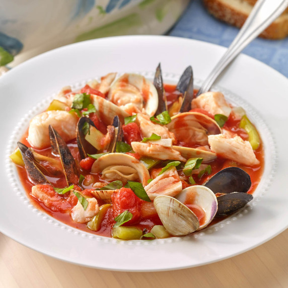 White bowl holding a portion of Bouillabaisse with soup tureen and sliced bread in the back