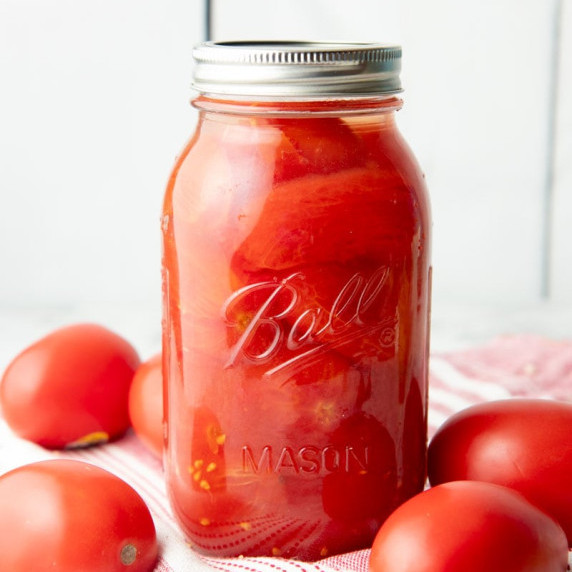 A jar of canned whole tomatoes stands on a red and white kitchen linen surrounded by fresh tomatoes.