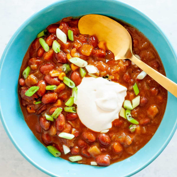 A gold spoon rests in a bowl of vegan chili topped with dairy-free cashew sour cream and scallions.