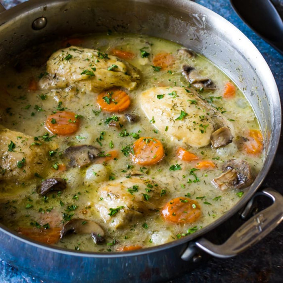 Overhead side view of chicken fricassee in a pot garnished with herbs on a blue surface.
