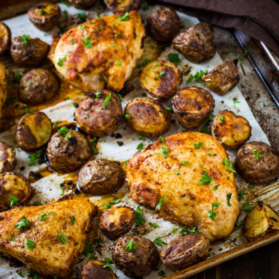 Side shot of chicken tray bake on a baking sheet lined with parchment paper garnished with herbs.