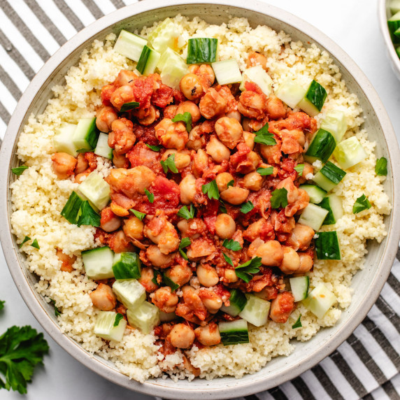 A bowl with tomato stewed chickpeas on top of couscous and diced cucumbers.