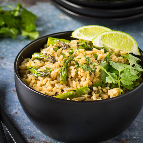 Side shot of rice with asparagus garnished with cilantro and lime wedges in a black bowl with forks.