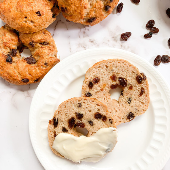 Sliced open cinnamon raisin bagel with cream cheese smear on a white plate with 3 bagels in top left