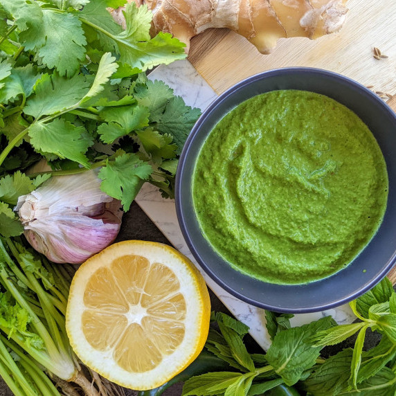 Green chutney in a black bowl on a wooden board, surrounded by herbs, lemon, ginger & garlic