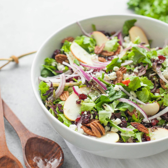 Cranberry, apple, and pecan salad in a white bowl with wooden serving spoons beside it.