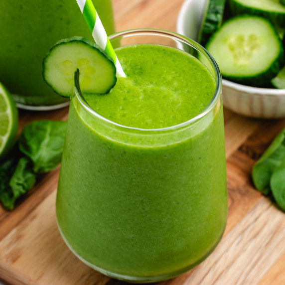 A green spinach and cucumber smoothie in a glass with a straw and a cucumber slice.