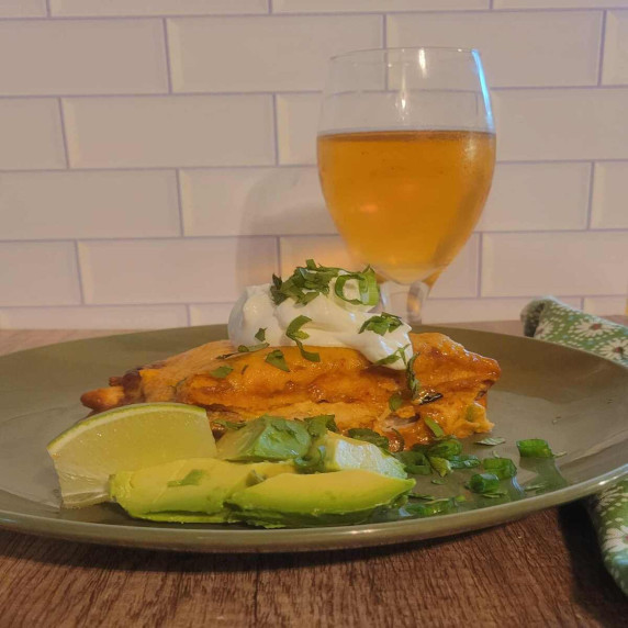 chicken enchilada casserole on a plate with lime, avocado, sour cream, green onion, and a cold beer