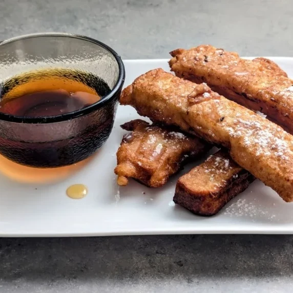 Four French Toast sticks stacked on a plate with a small bowl of maple syrup for dipping. 