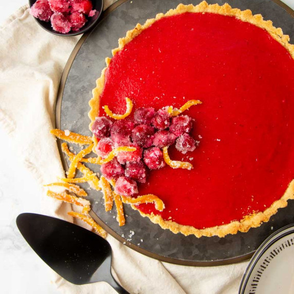 Cranberry curd tart garnished with sugared cranberries and candied orange peels on a serving tray.