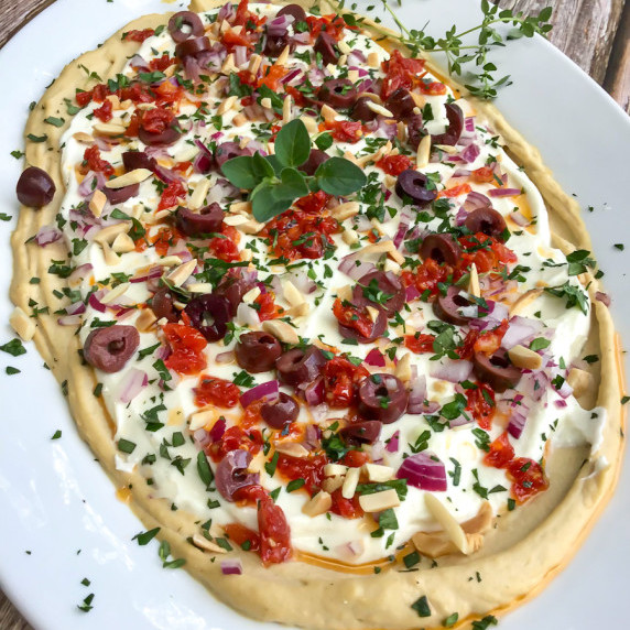 a layer of hummus topped with lemon whipped feta, fresh herbs, sun dried tomatoes, olives, almonds