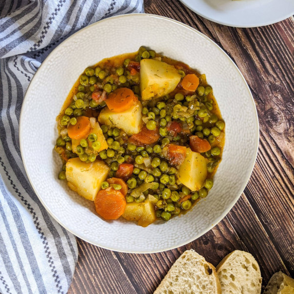 Stewed green peas served in a white bowl next to bread and feta cheese.
