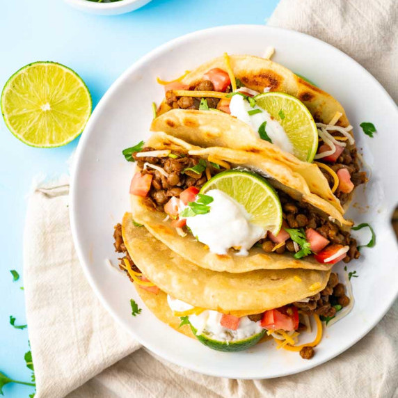 Overhead of three lentil tacos on a plate garnished with fresh lime, sour cream, and diced tomatoes.