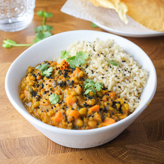photo of a white bowl with brown rice and mung bean curry and poppadoms in the background