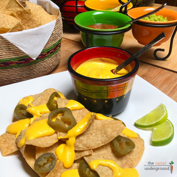 A plate of nachos with vegan cheese sauce, sprinkle with sliced jalapeños and a side of lime slices.