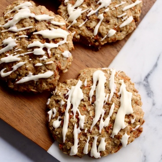 oatmeal carrot cake cookies are drizzled with cream cheese glaze and served on a marble and wooden b