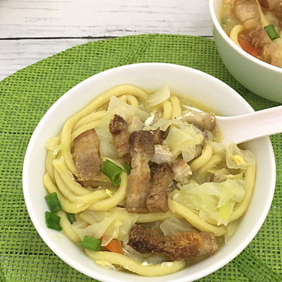 pancit lomi or egg noodle soup served in a clear large bowl with soup spoon beside it