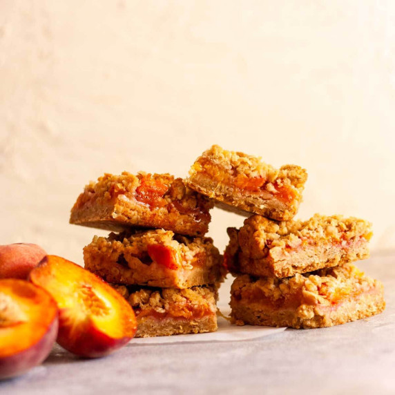 Stacked peach crumble bars on a gray and white surface next to sliced peaches.