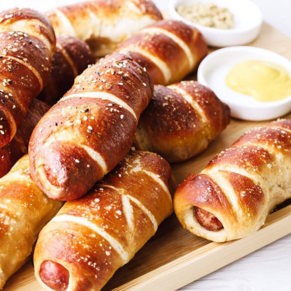 Pretzel dogs stacked on a wooden block. There are 2 dipping sauces in white bowls.