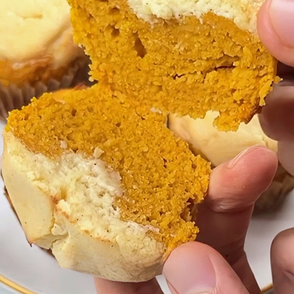 A hand breaks open a keto pumpkin muffin, revealing a layered interior of yellow cake and white fill