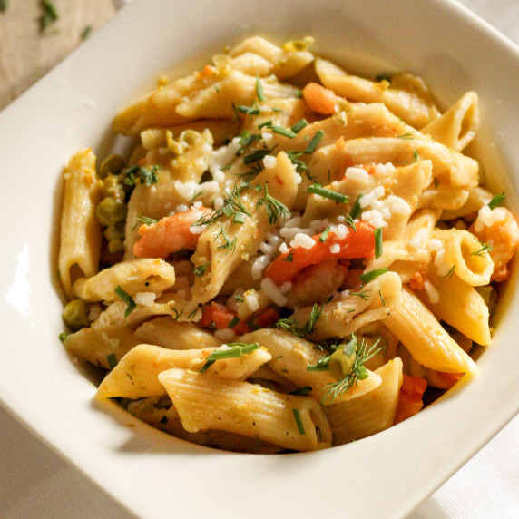 A white bowl with rigatoni pasta, red bell peppers, chives, dill and fresh tomato sauce, shrimps
