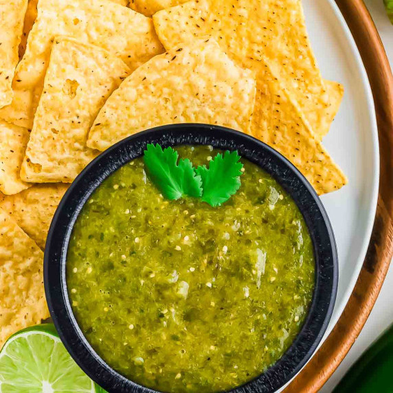 A small bowl of green salsa verde is surrounded by tortilla chips on a white plate.