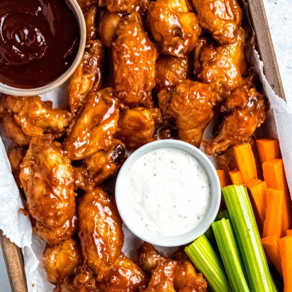 Oven baked chicken wings in barbecue sauce and served on a parchment lined wooden tray with veggies.