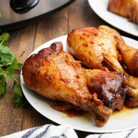 Four chicken drumsticks resting in flavorful juices on a white plate with fresh parsley alongside.