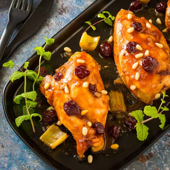 Overhead shot of three pieces of cranberry chicken garnished with cranberries and herbs on a black p