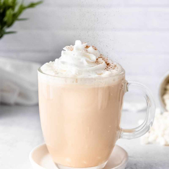 Cinnamon sprinkles down onto the whipped cream garnish on a white chocolate mocha in a glass mug.