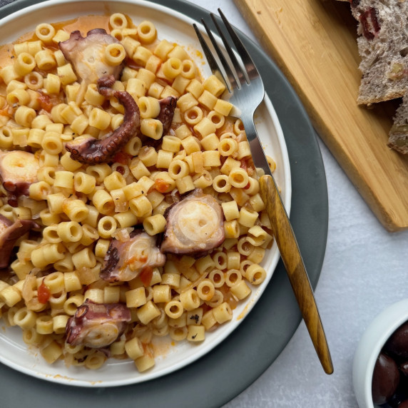 Octopus pasta served on white plate with bread slices and olives in the background.