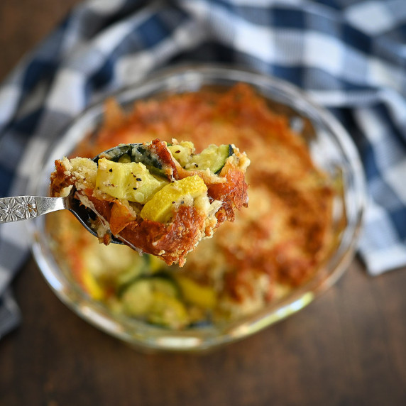 A spoonful of cheesy squash being served from a clear baking dish. 