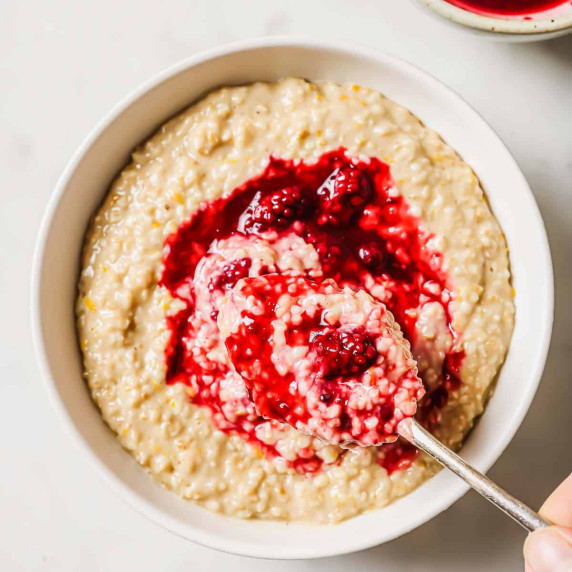 A bowl of steel-cut oatmeal topped with blackberry sauce. A spoon is taking a spoonful of oatmeal.