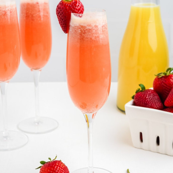 A champagne flute with fizzy pink strawberry mimosa stands with additional mimosas and fresh berries