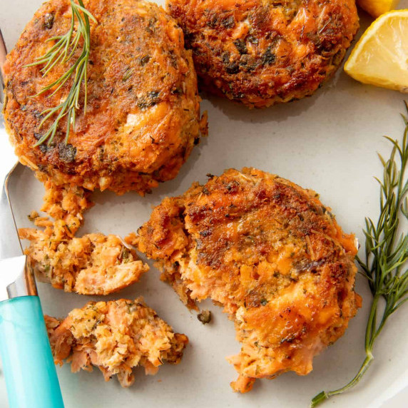 Close up of three salmon cakes on a plate with a fork, fresh dill, and lemon slices.