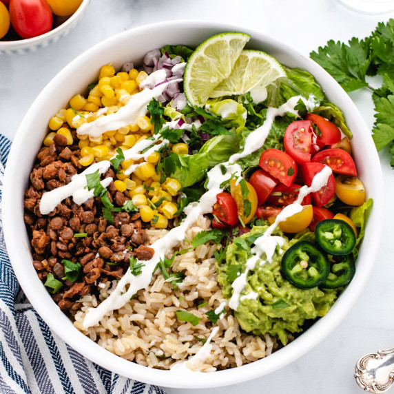 Taco bowl with corn, lentil taco meat, rice, guacamole, cherry tomatoes, and lime slices.