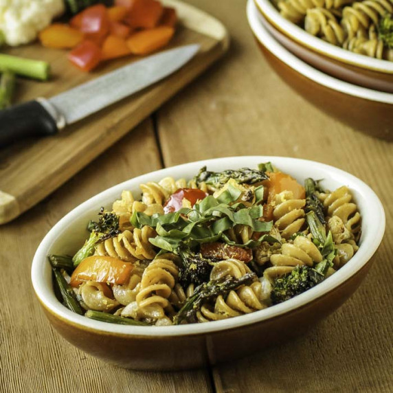 Side shot of veggie rotini in an orange and white oval bowl with chopped veggies on a wooden cutting